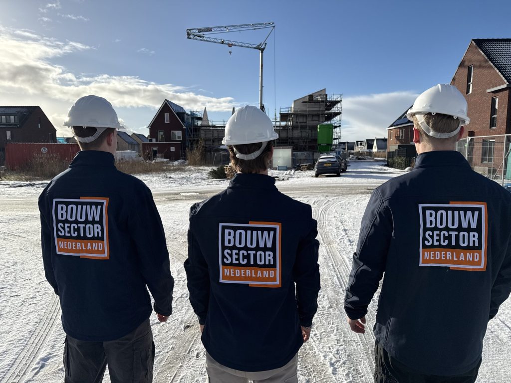 Drie vakmensen van Bouwsector Nederland met bouwhelm op een besneeuwde straat langs bakstenen woningen