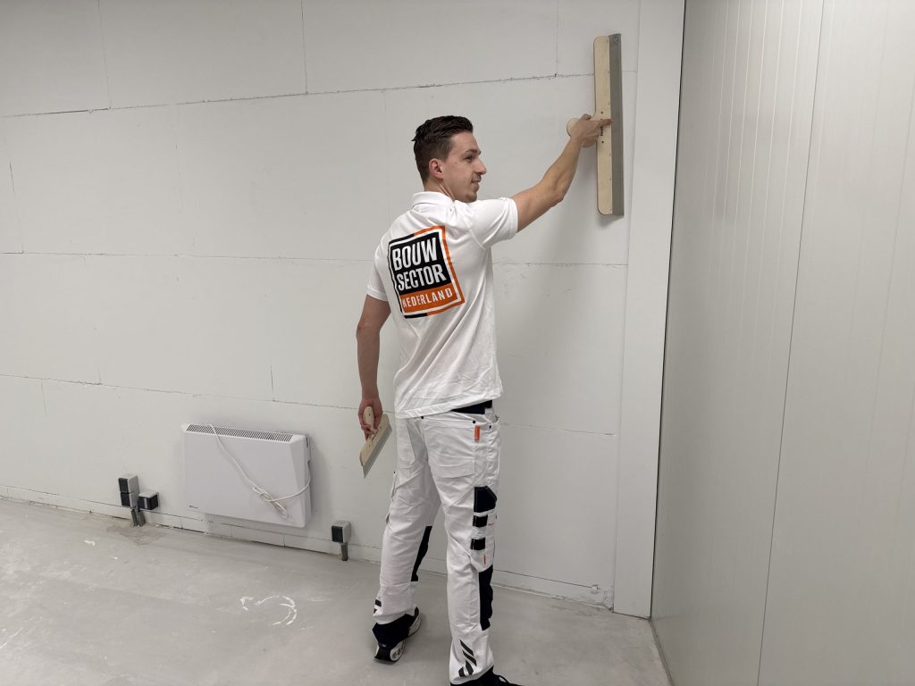 Stukadoor kijkt opzij terwijl hij een lange spaan hoog tegen een kale wand houdt