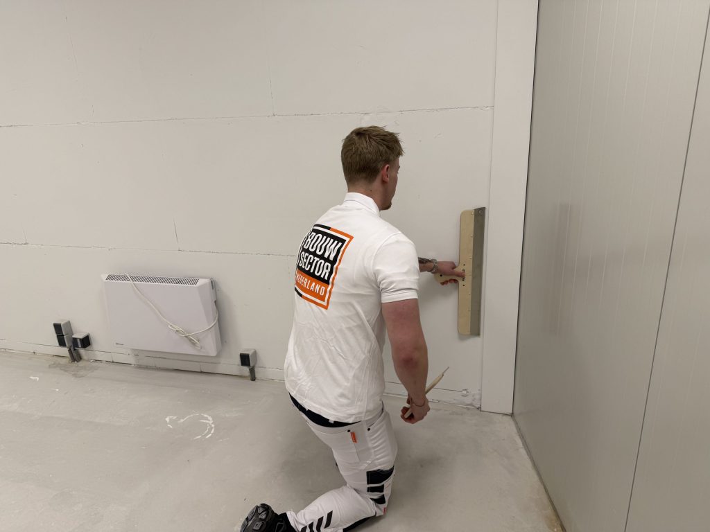 Geknielde stukadoor werkt met een spaan het onderste deel van een wand bij, naast een radiator